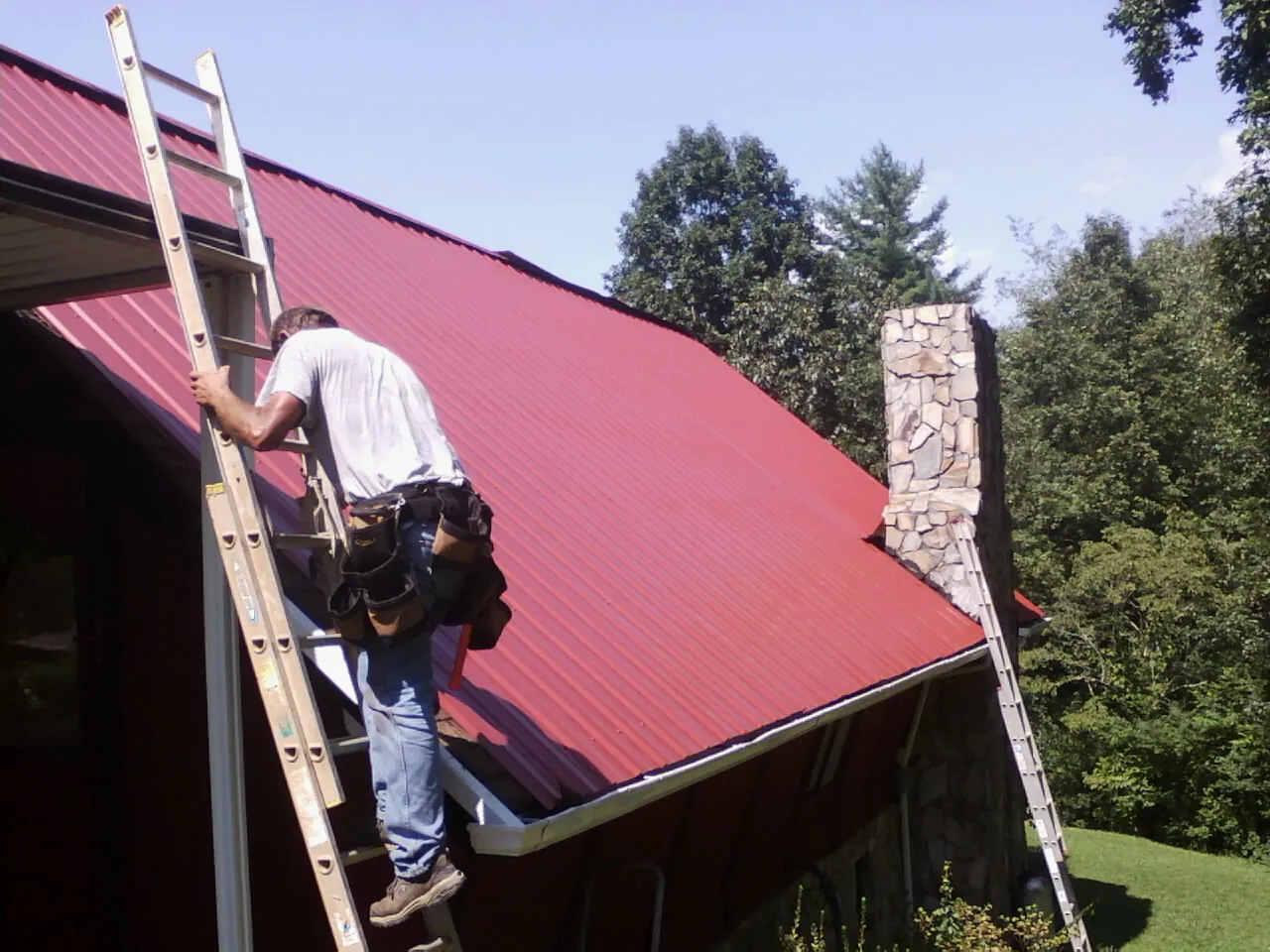 Roofer on a ladder beside a red metal roof and stone chimney — Soffit & Fascia Repair by Roofing Brothers in Basehor
